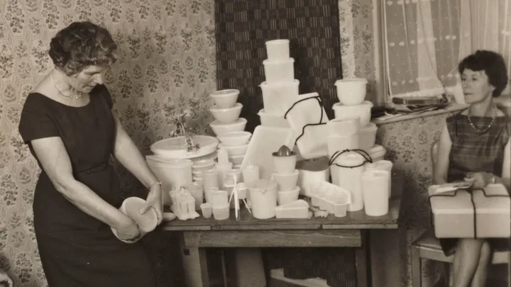 Two women in vintage attire at a Tupperware party; one demonstrates products, the other holds a boxed set, circa 1950s.