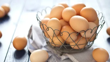 A wire basket filled with brown eggs sits on a striped cloth, with a few eggs scattered on a blue wooden table.