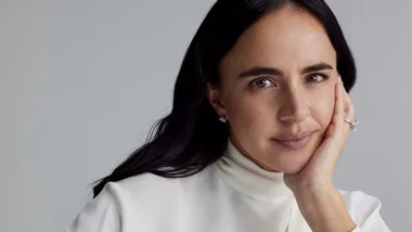 A woman with dark hair and a white top rests her chin on her hand, wearing a ring and earrings, against a gray background.