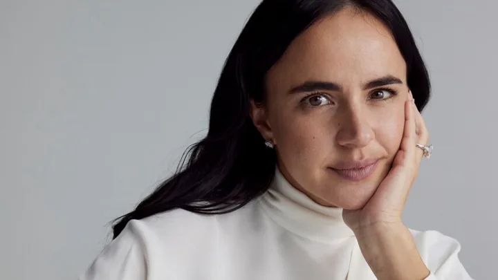 A woman with dark hair and a white top rests her chin on her hand, wearing a ring and earrings, against a gray background.