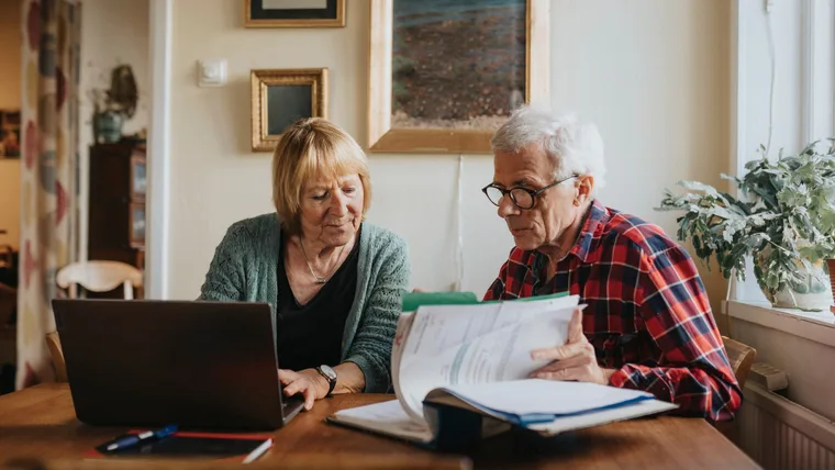 Elderly couple reviewing documents at a table, with a laptop and indoor plants in the background.