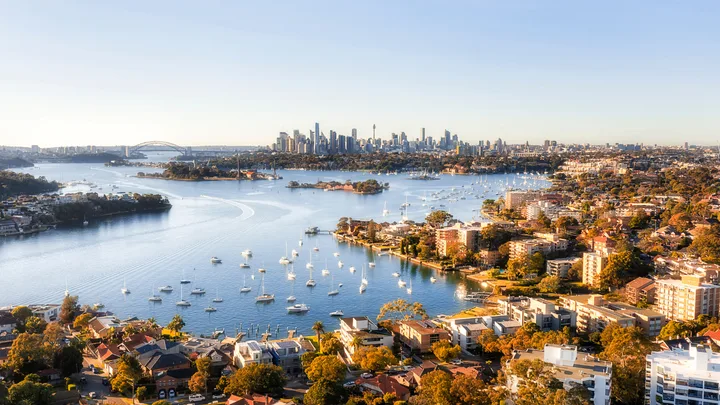 Aerial view of Sydney Harbour with boats, residential areas, and city skyline, including the Sydney Harbour Bridge.