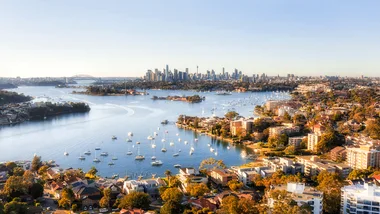 Aerial view of Sydney Harbour with boats, residential areas, and city skyline, including the Sydney Harbour Bridge.