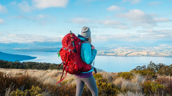 A hiker with a red backpack overlooks a scenic lake and mountains under a blue sky.