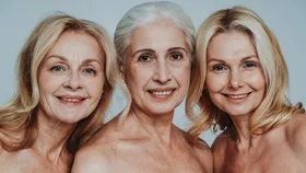 Three mature women with fair hair and bare shoulders smiling against a light blue background.