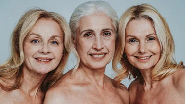 Three mature women with fair hair and bare shoulders smiling against a light blue background.