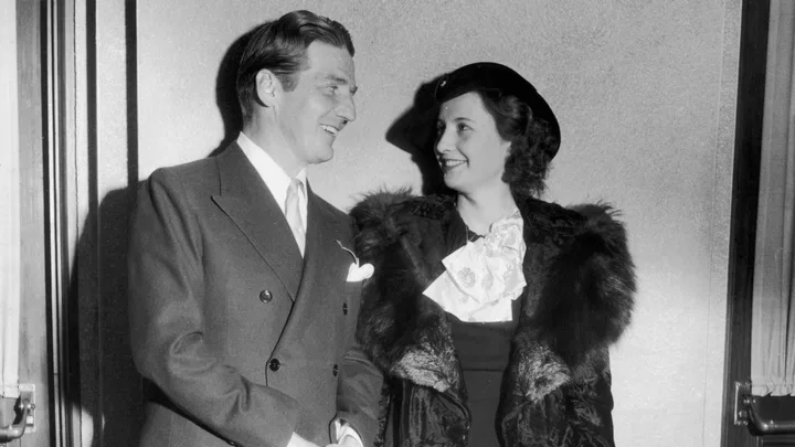 A man in a suit and woman in a fur-trimmed coat, chatting and smiling in a classic black and white photo.