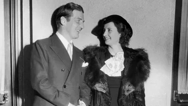 A man in a suit and woman in a fur-trimmed coat, chatting and smiling in a classic black and white photo.