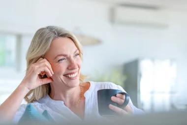 Mature woman enjoying a cup of tea or coffee sitting on the sofa. She is happy and smiling looking joyful.