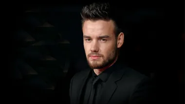 A man in a black suit with a beard and styled hair against a dark background.