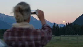 A person taking a photo of a moonrise over a mountain landscape with a smartphone.