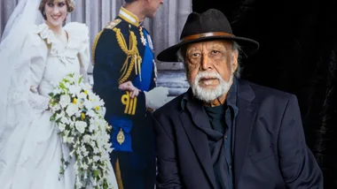 Man in a hat and suit in front of a photograph of a bride and groom, possibly a royal wedding scene.