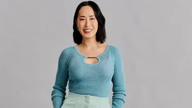 A person smiling, wearing a blue sweater with a cut-out, standing against a gray background.