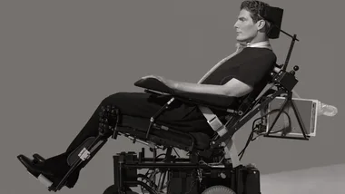 Man in reclining wheelchair-like device, wearing dark clothing, against a gray background.