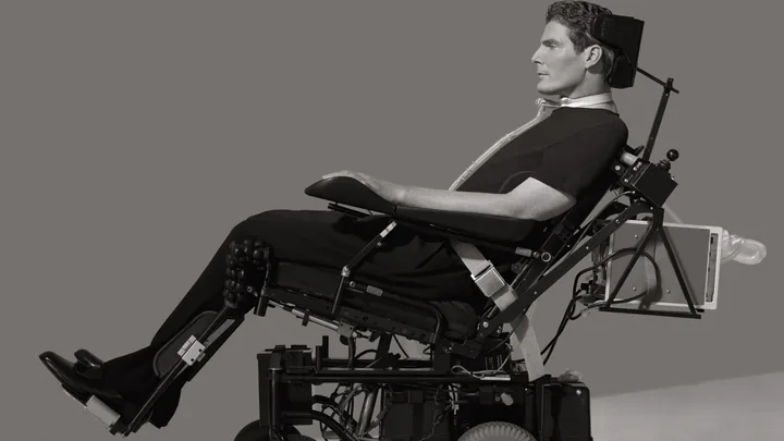 Man in reclining wheelchair-like device, wearing dark clothing, against a gray background.