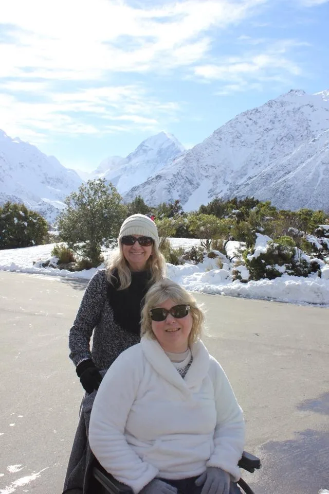 Dee stands behind Lee-Ann's wheelchair which is parked in front of three spectacular snow-capped mountain peaks.