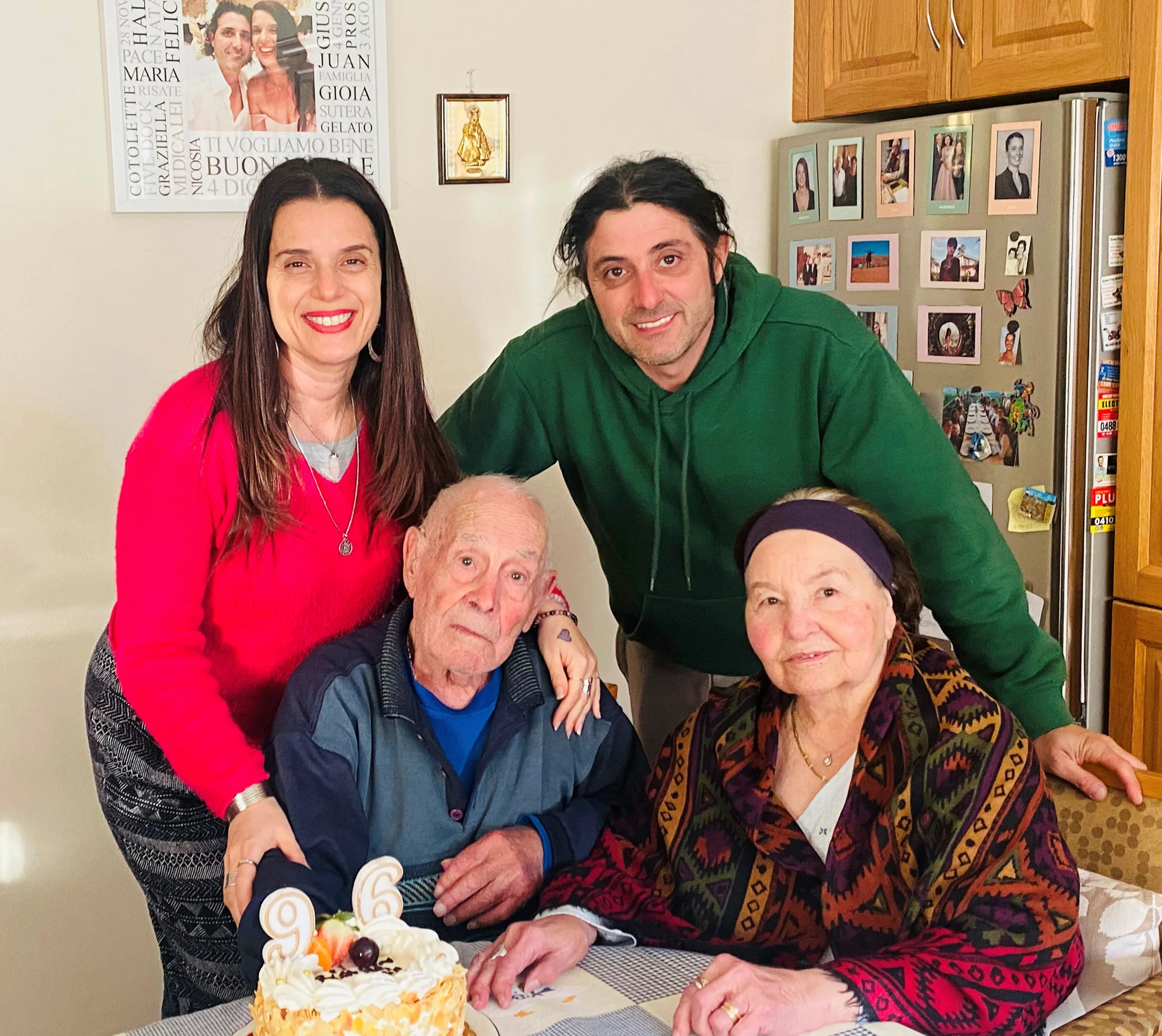 Maria, wearing a red jumper, at left, in a group photograph with her partner and her parents. The photograph is taken at their kitchen table.