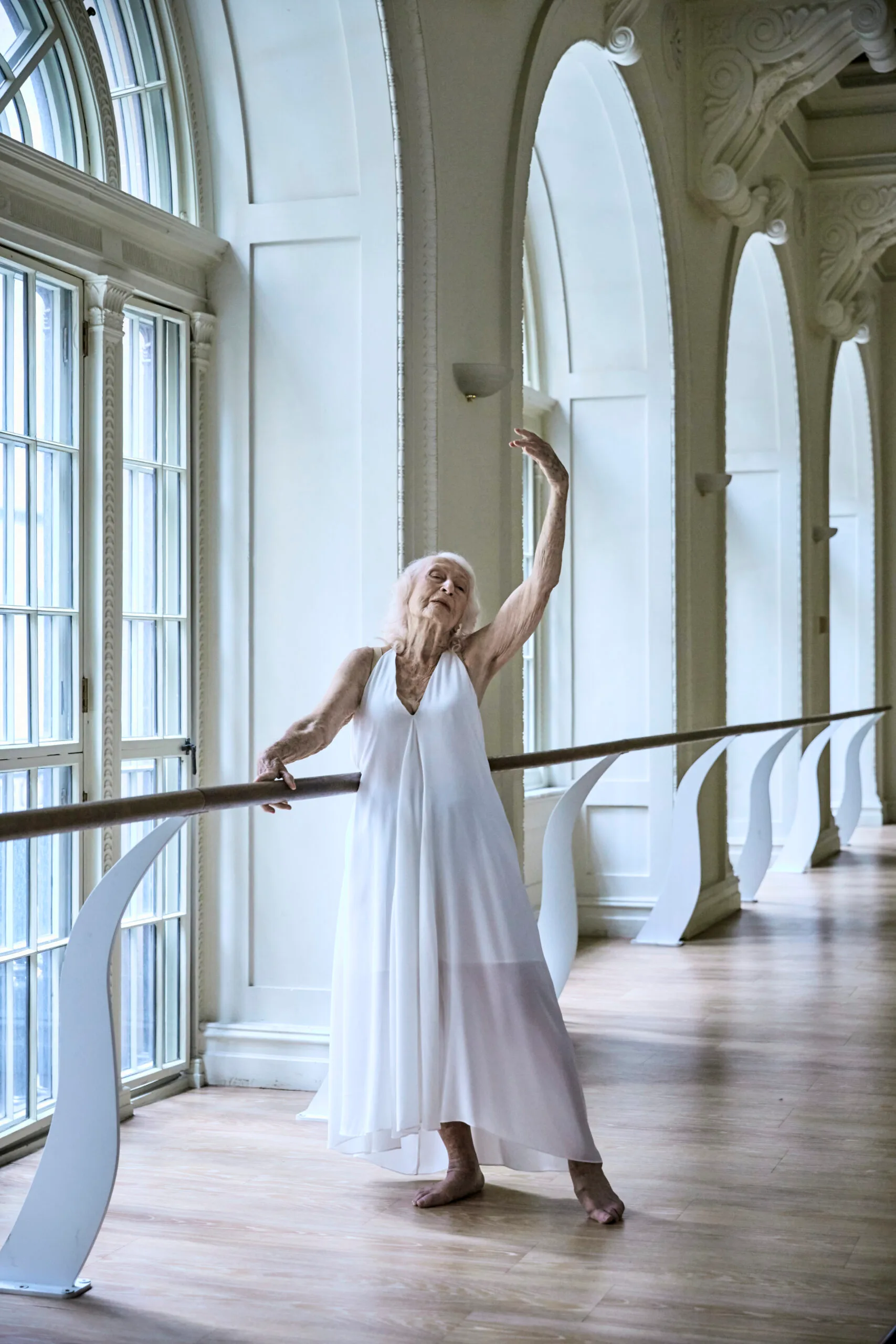 Eileen wears a white shift dress and demonstrates ballet exercises at barre in a sun drenched studio.