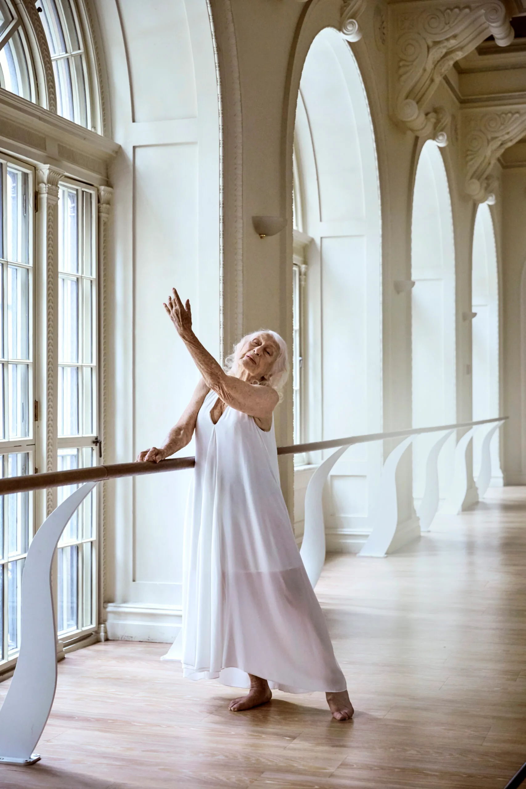 Eileen dressed in a long white dress demonstrates a ballet exercise at barre in a beautiful light-filled studio.