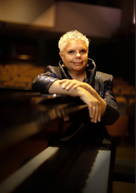 Deborah Cheethham Fraillon, her hair short and grey, sits in a dark auditorium and smiles. She is wearing a grey sparkly jacket.