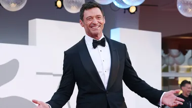 Man in a tuxedo smiling at an event with a red carpet backdrop.