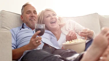 Middle-aged couple laughing on a sofa, enjoying popcorn and wine while watching TV.