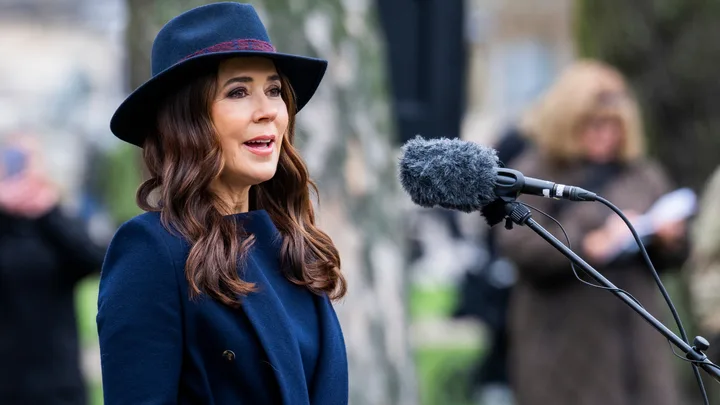 A woman wearing a navy coat and hat speaks into a microphone outdoors.