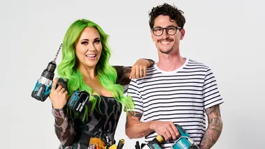 Two individuals smiling, hold power tools; one has vibrant green hair, the other wears glasses and a striped shirt.