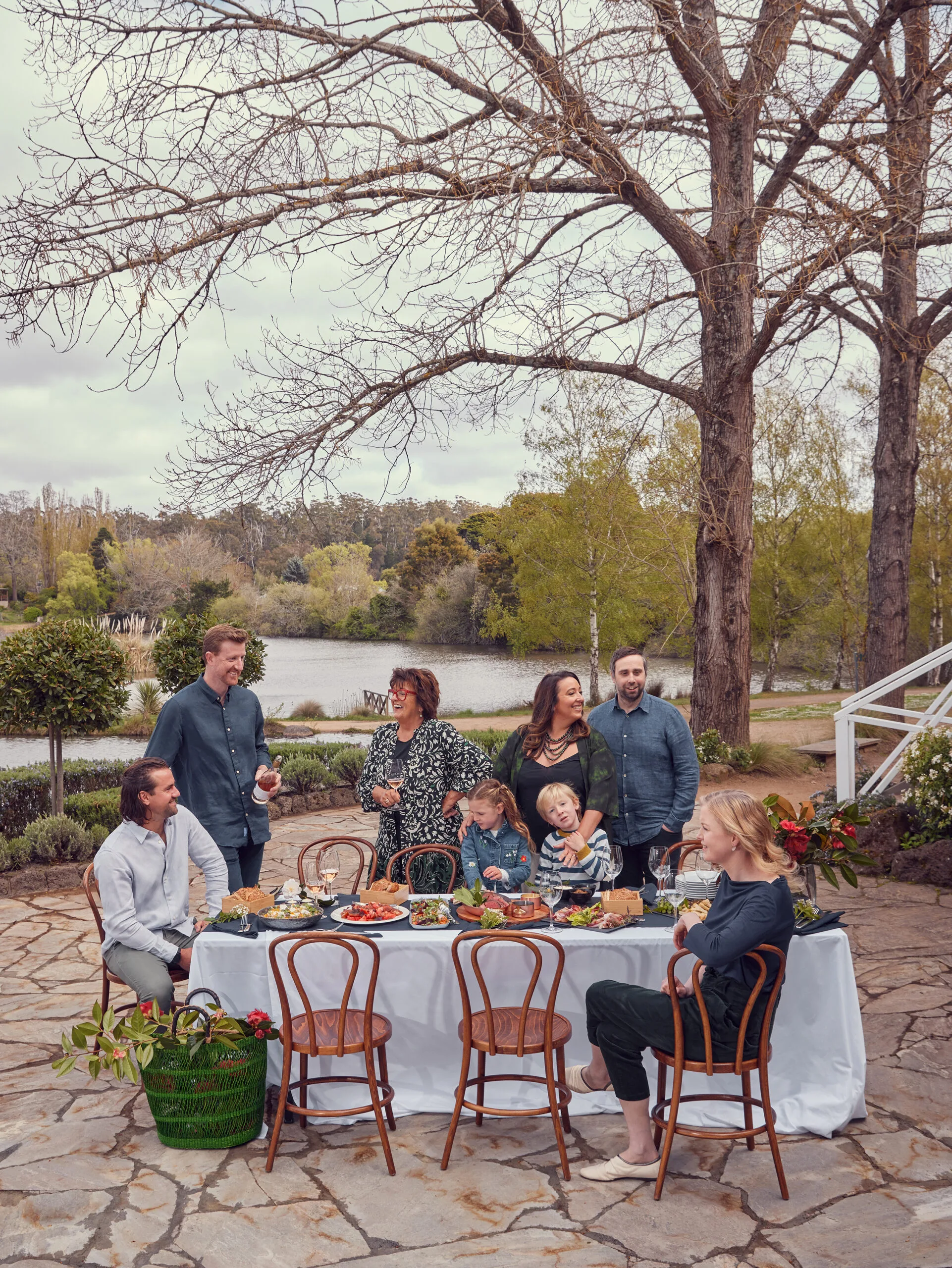 Alla Wolf-Tasker and her family friends gather around a table laden with food. In the background the lake is surrounded by trees.