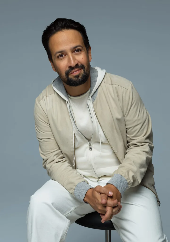 Lin-Maneul Miranda dressed in all white with a beige jacket. Sits on a stool.