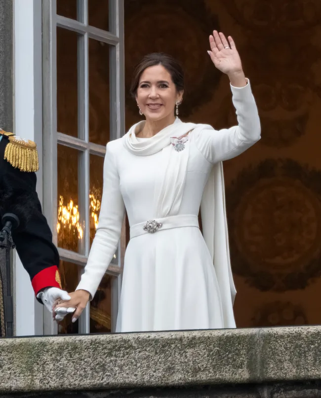 A woman in a white dress waves while standing on a balcony, holding hands with a person in a formal black and red uniform.