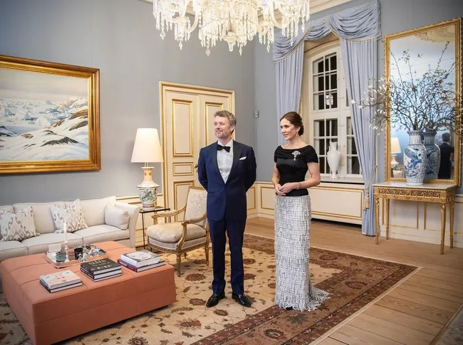 Elegant room with chandelier, art, and two people dressed formally; sitting area features decorative elements.