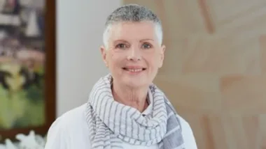 Elderly woman smiling, wearing a striped scarf and white top, with short gray hair, standing indoors.