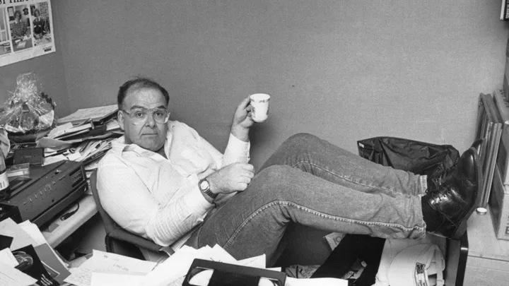 Man in glasses with feet on desk, holding a cup, surrounded by cluttered papers and office equipment.