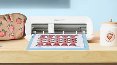 Cricut Joy Xtra cutting machine on table with strawberry designs being cut, next to a pouch and jar with similar designs.