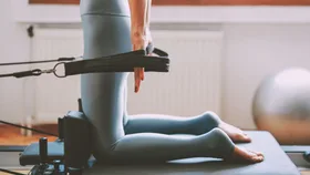 Woman in blue tights performing a reformer Pilates workout, pulling bands behind her back with bare feet. The gym setting includes an exercise ball in the background.