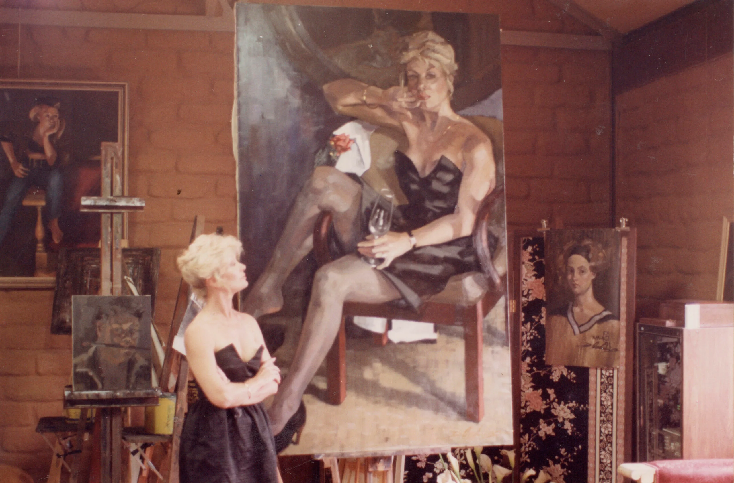 A young Di Morrissey, a white woman with blonde hair, stands by a large portrait of herself.