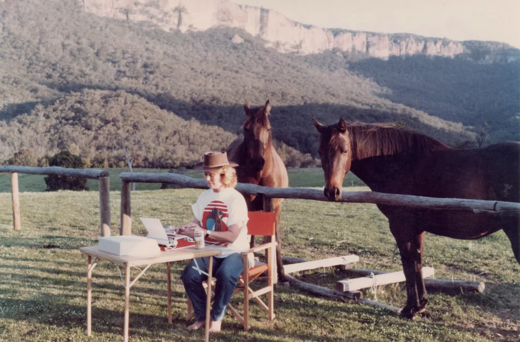 Di Morrissey at a card table writing on a laptop. Behind her are two horses, and in the background are green hills.