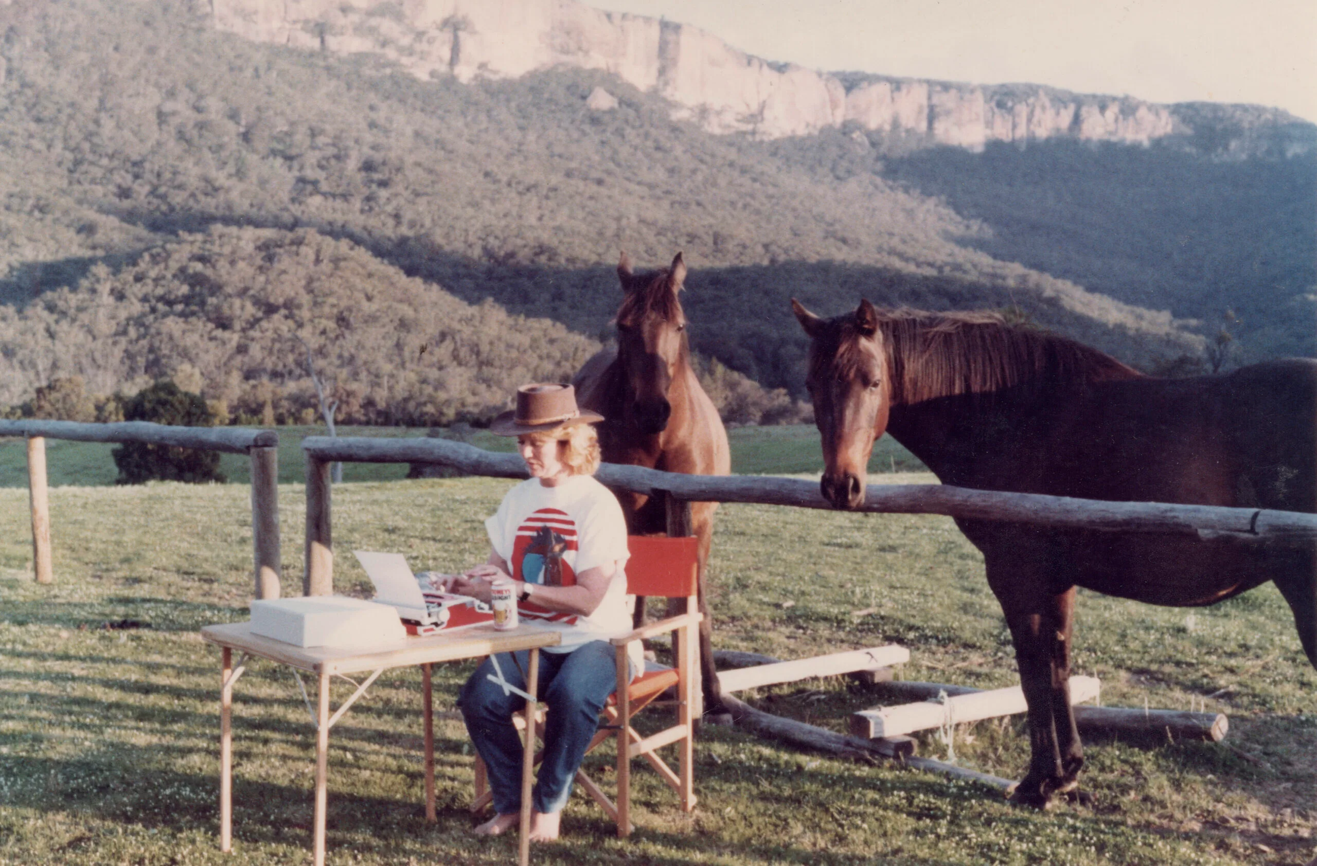 Di Morrissey at a card table writing on a laptop. Behind her are two horses, and in the background are green hills.
