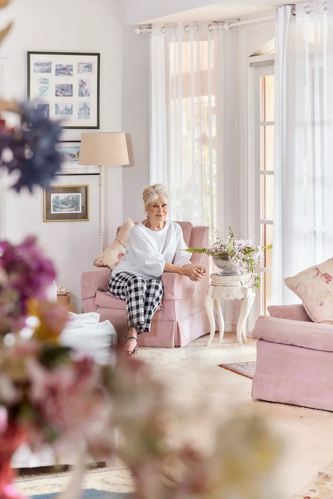 Author Di Morrissey, a white woman with blonde hair, sits on a pink lounge chair in a living room.
