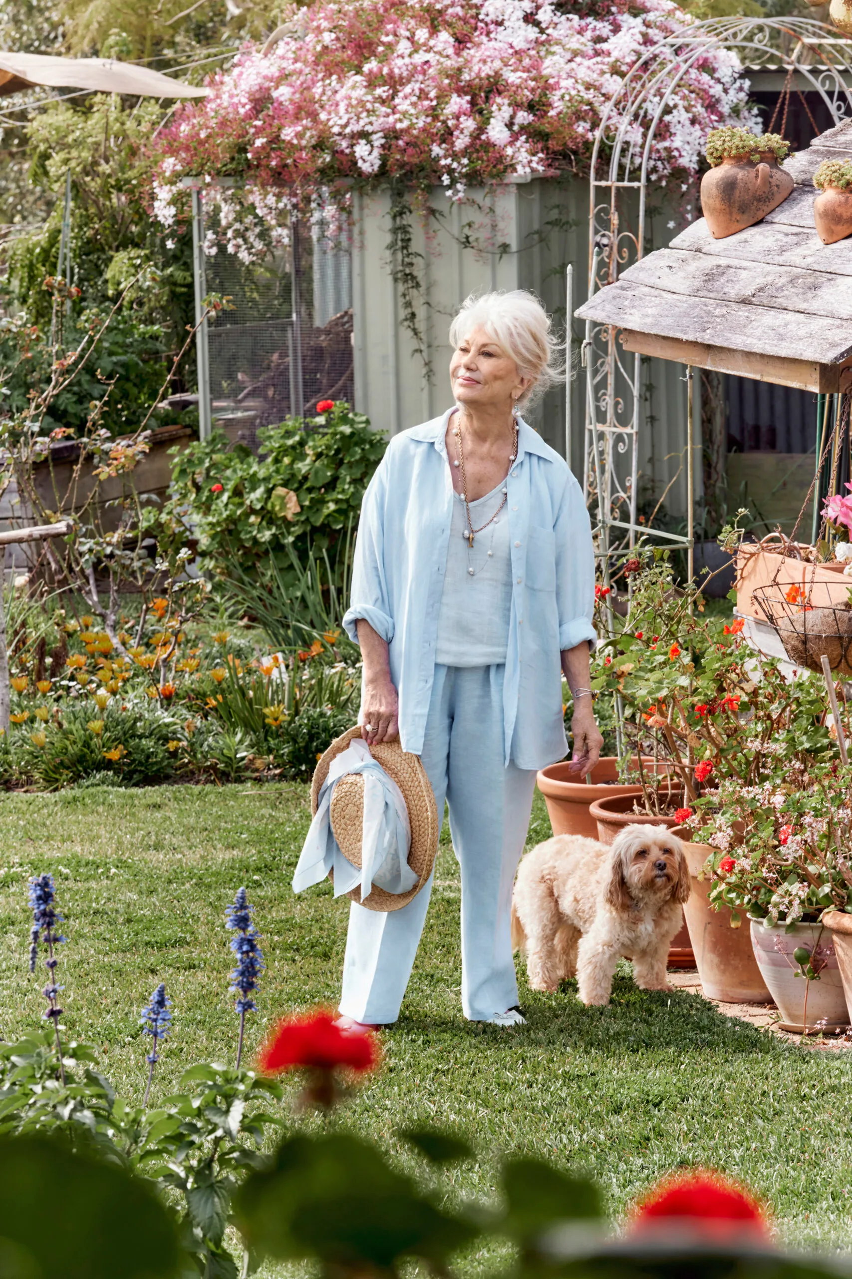 Author Di Morrissey, a white woman with blonde hair, in a garden with a small dog.