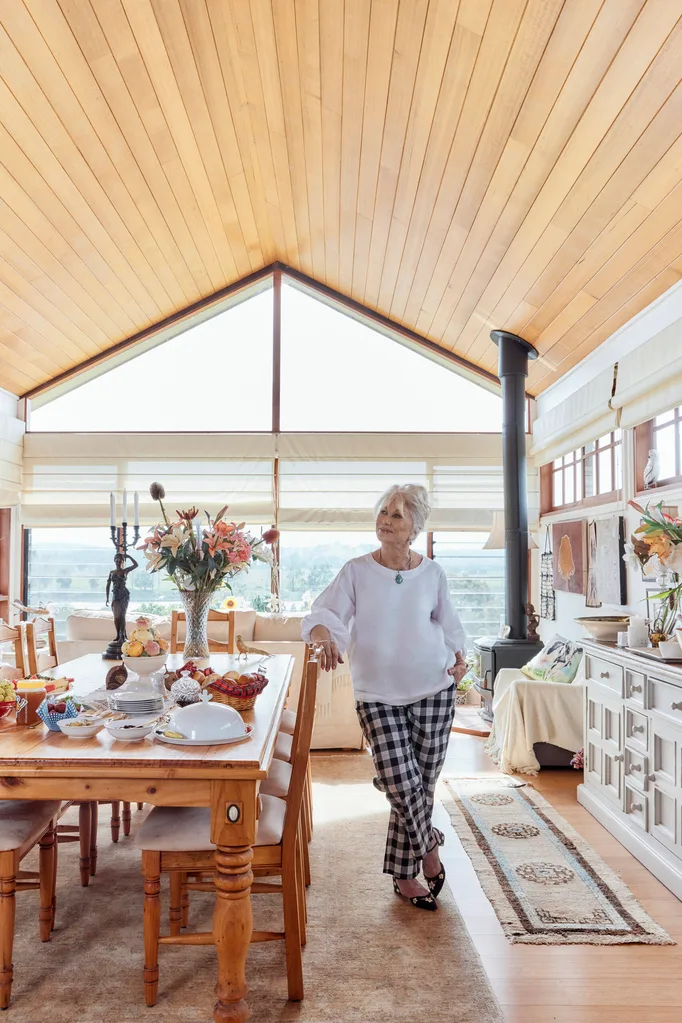 Author Di Morrissey, a white woman with blonde hair, stands in a living room by a large wooden table.