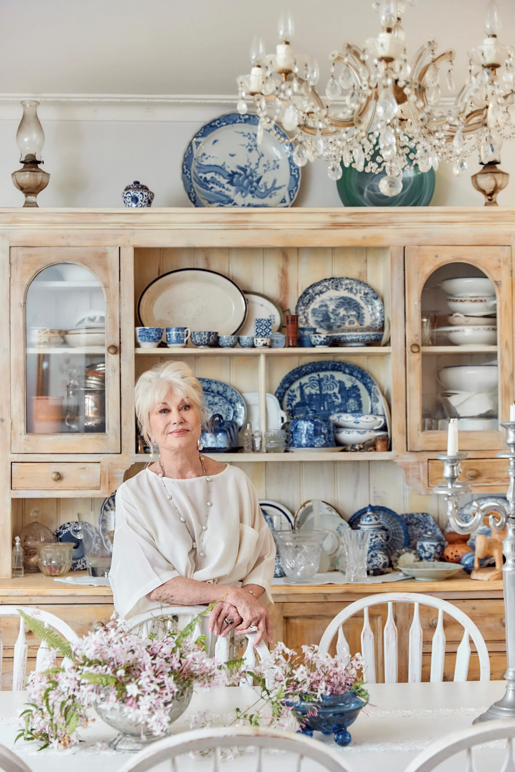 Author Di Morrissey, a white woman with blonde hair, stands in front of a cabinet filled with decorative crockery.