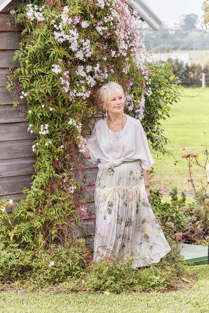 Author Di Morrissey, a white woman with blonde hair, stands by a flower-covered trellis.