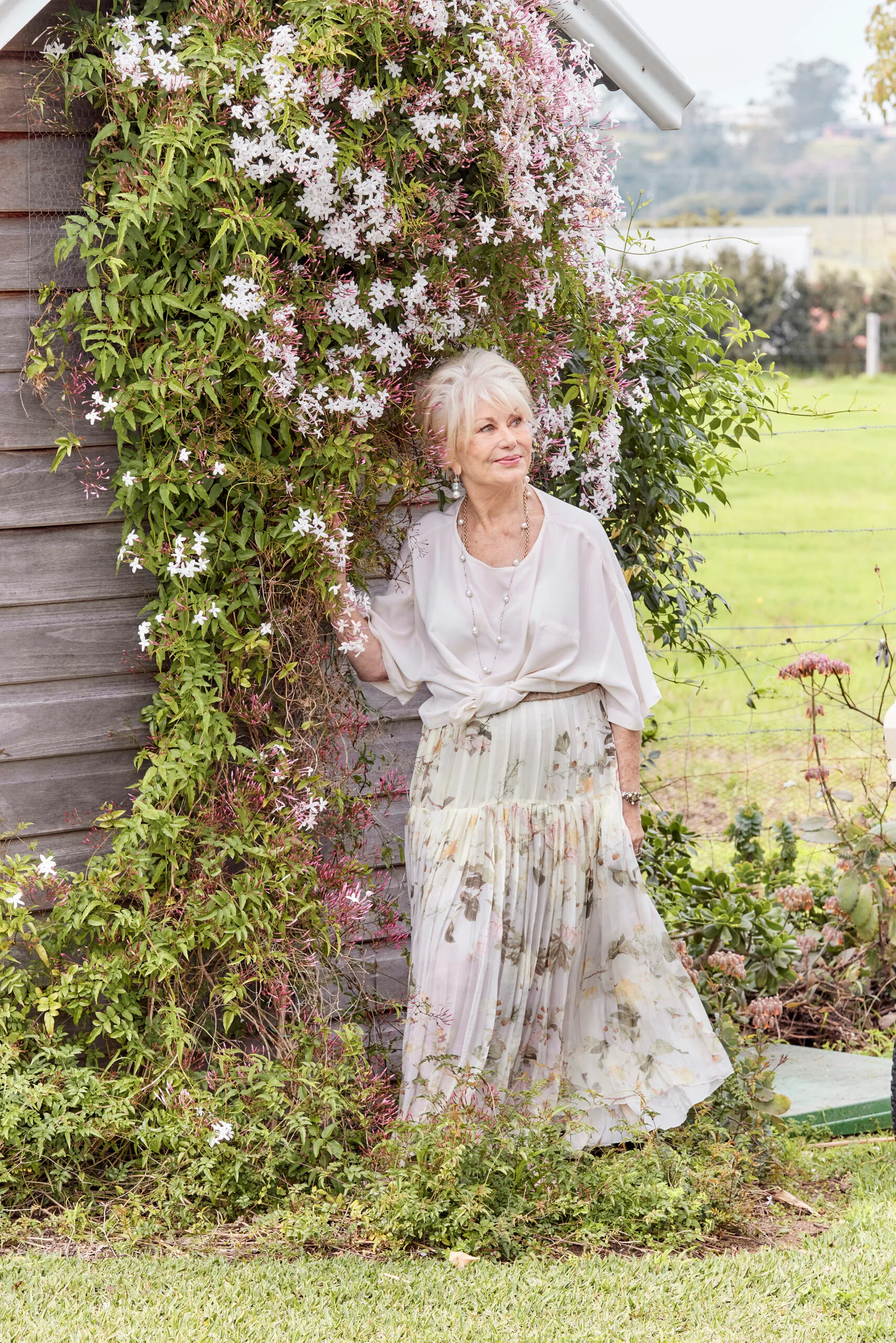 Author Di Morrissey, a white woman with blonde hair, stands by a flower-covered trellis.