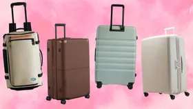 Four different styles of travel luggage arranged against a vibrant pink watercolour background. From left to right: a beige Bagsmart Blast 43L Roller Backpack with orange accents, a dark brown July Checked Plus Expandable suitcase, a mist-blue Antler Icon Stripe large suitcase, and a cloud-white Nere Bondi 80cm hardshell suitcase.