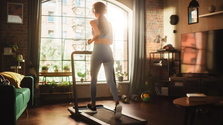 Woman Running on a Treadmill at Her Home Gym. Energetic Workout: Female Training while Listening Podcast or Music in Headphones. Apartment with Window View. Side Back View