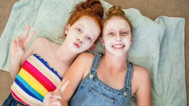 Two teen girls have facials with facial mask and cucumbers to eyes in a relaxed, fun home environment.