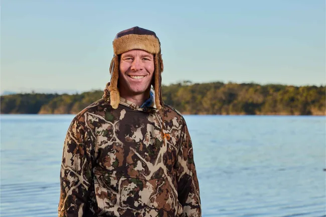 Man in camo hoodie and fur hat smiling by a lake with forest background.