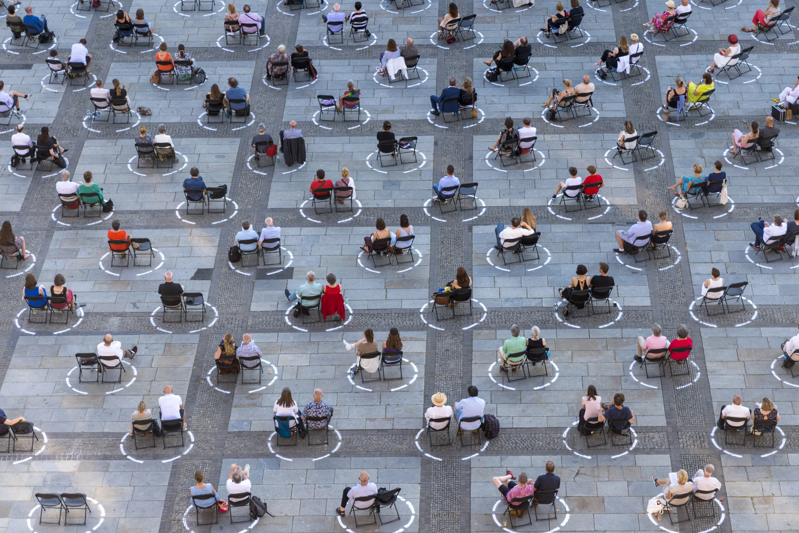 People sit on chairs in groups of two within circles that have been drawn on a grey outdoor area.